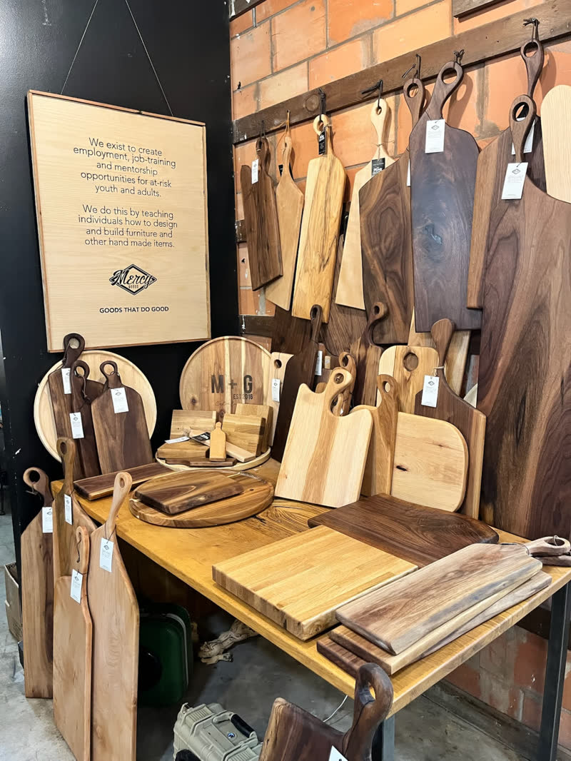 A display of handmade cutting and serving boards beneath the Mercy Goods mission sign