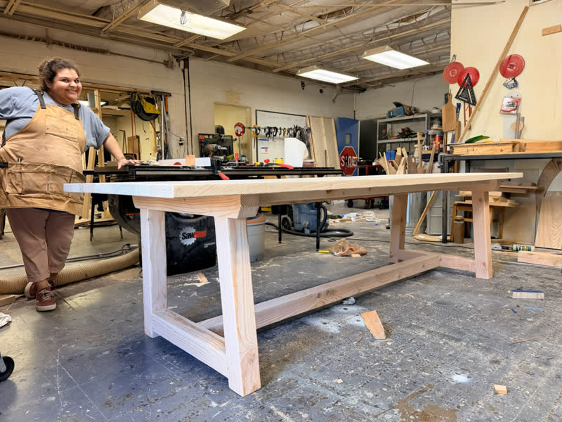 A young maker in a shop apron standing proudly beside a long table she built