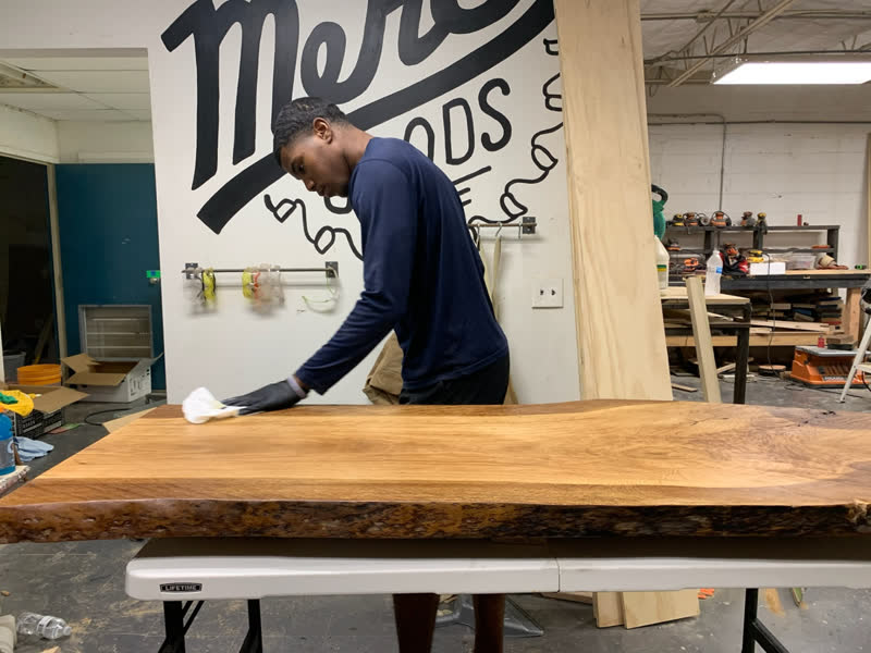 A young maker hand-rubbing finish into a live-edge slab beneath the Mercy Goods mural