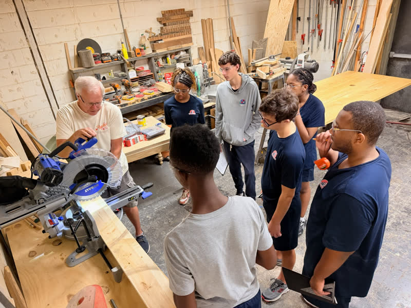 A mentor demonstrating the miter saw to a group of young makers gathered around the bench