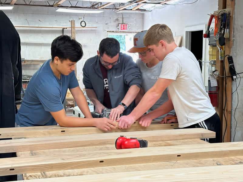 A mentor and three young makers fitting pocket-hole rails for a tabletop, ear muffs waiting on the bench