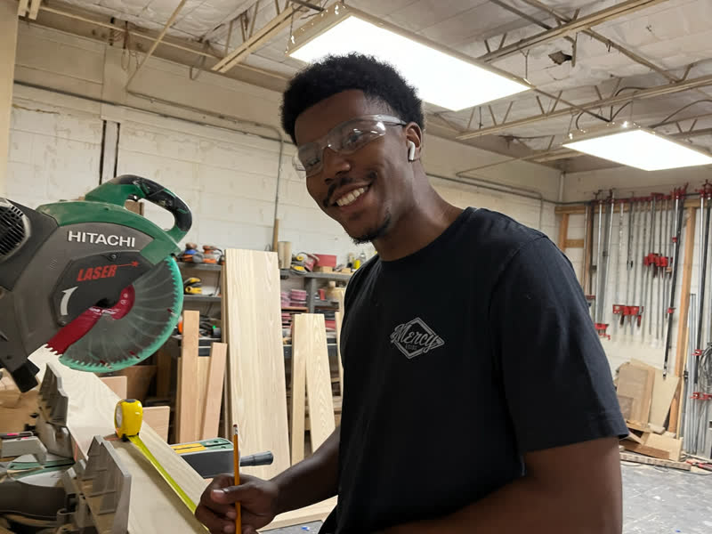 A young maker in a black Mercy Goods shirt smiling at the miter saw