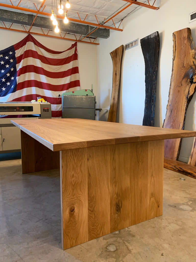 A thick white oak table on a solid plank base, in front of the flag and raw slabs