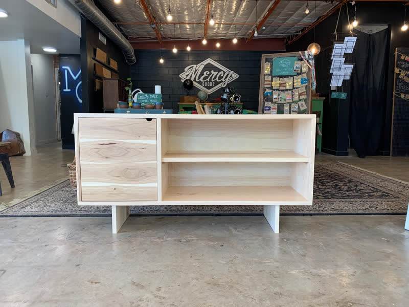 A light wood credenza with cabinet doors beneath the painted Mercy Goods sign