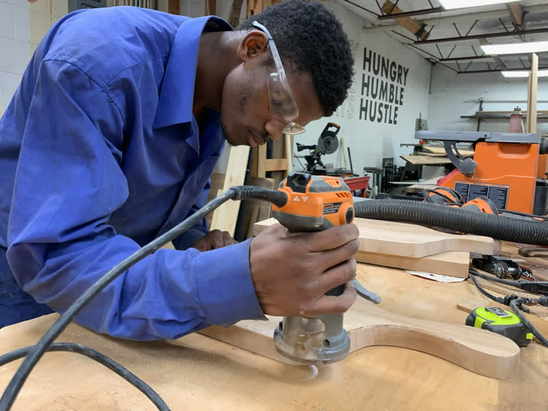 A young maker in safety glasses routing the edge of a serving board under the Hungry Humble Hustle wall