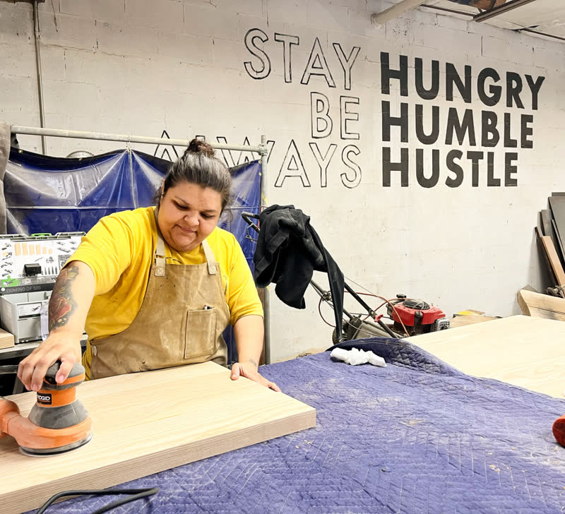 A young maker sanding a wide board beneath the Stay Hungry, Be Humble, Always Hustle mural