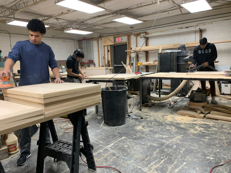 Three young makers sanding stacks of tabletops across the workshop