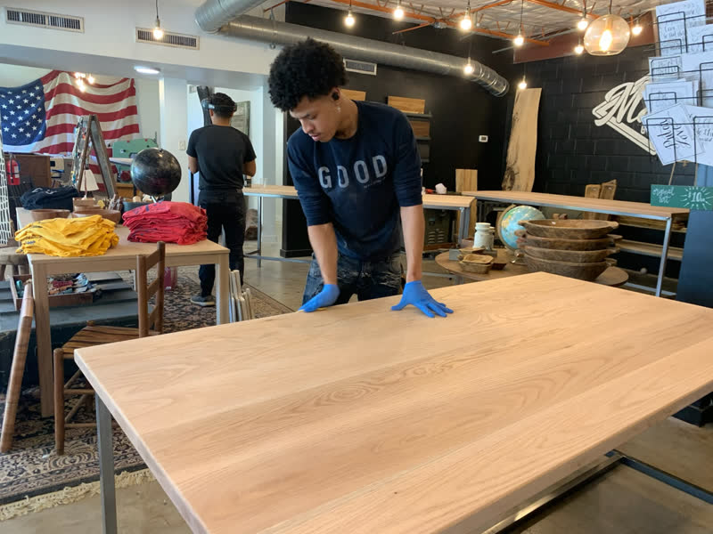 A young maker hand-finishing an oak tabletop with blue gloves in the Mercy Goods shop