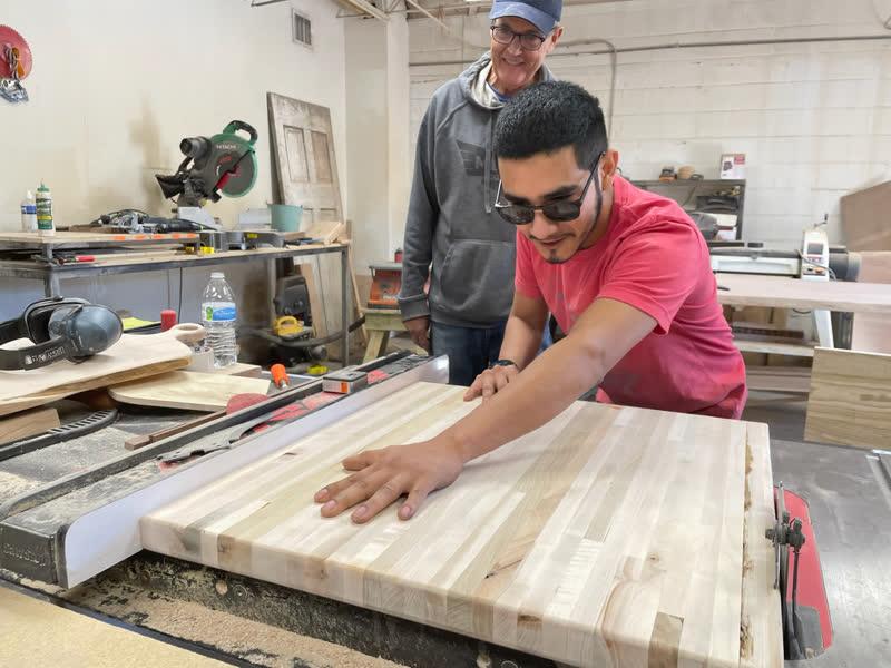A young maker trimming a butcher block top at the table saw while a mentor watches