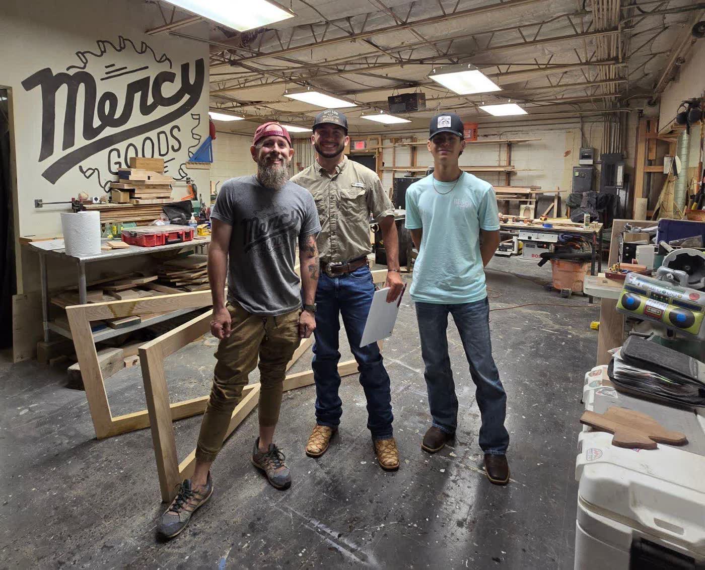 A mentor and two students standing together in the Mercy Goods workshop beneath the mural