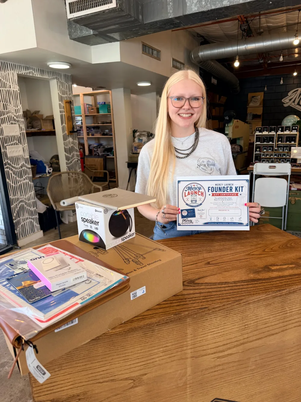 A smiling young woman holding her Mercy Launch Founder Kit certificate in the Mercy Goods shop