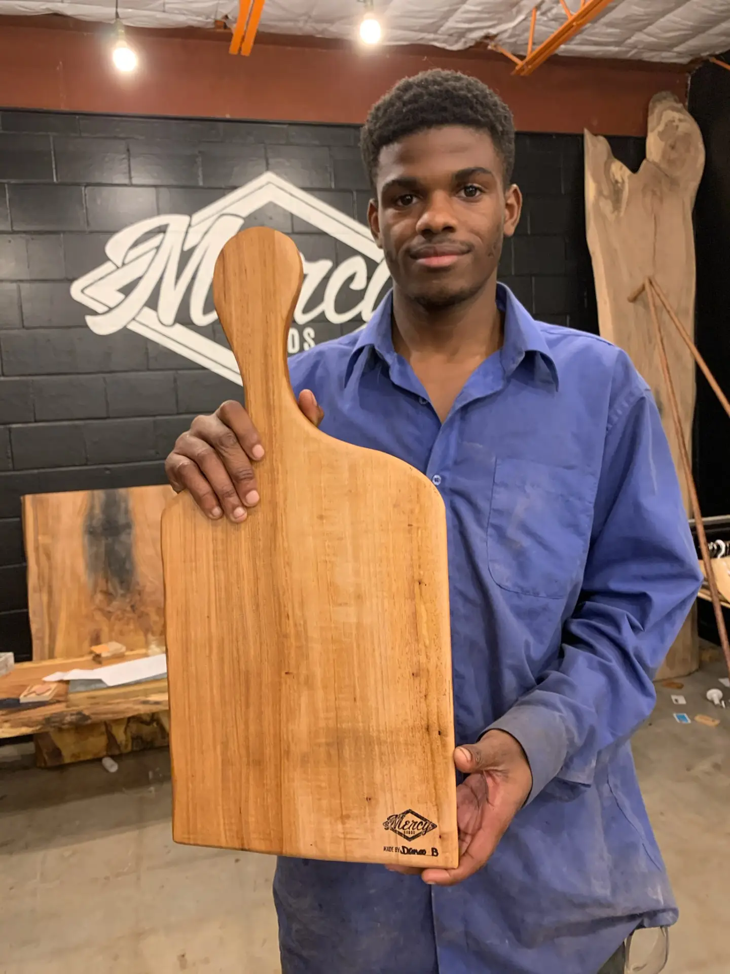 A young maker holding his finished serving board in front of the Mercy Goods mural