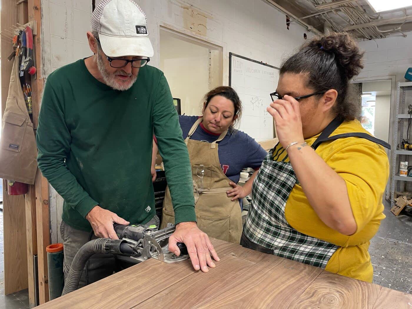 A mentor teaching two Makers to use a router on a walnut tabletop in the Mercy Goods workshop
